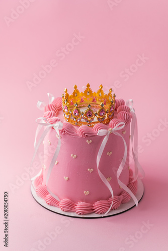 Pink princess birthday cake with golden crown topper, ribbon bows and heart details on pastel pink background. Elegant celebration dessert for girls party or baby shower