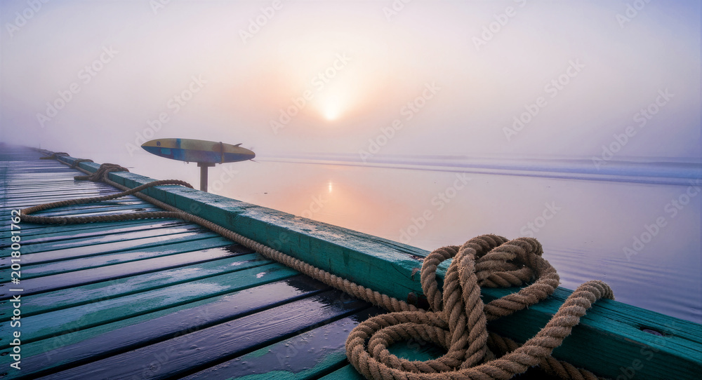 Fototapeta premium Nautical ropes resting on a wooden pier overlooking a misty beach at sunrise. Soft morning light illuminates the fog over calm ocean waves. Coastal serenity concept, ideal for travel marketing.