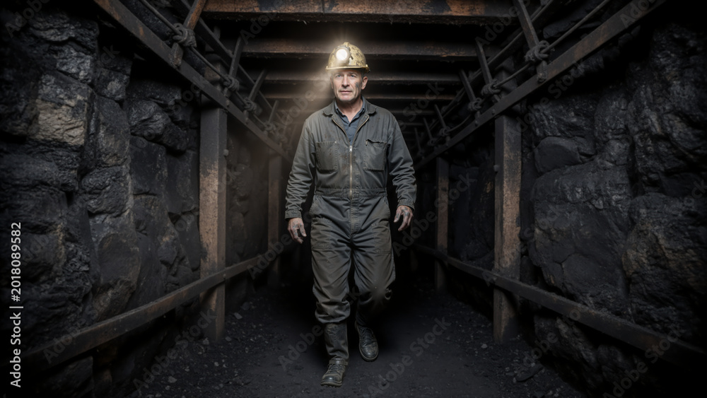 Fototapeta premium Middle aged male coal miner walking in a dark underground mine shaft. Industrial worker wearing a dirty jumpsuit and hard hat with illuminated headlamp. Heavy manual labor concept