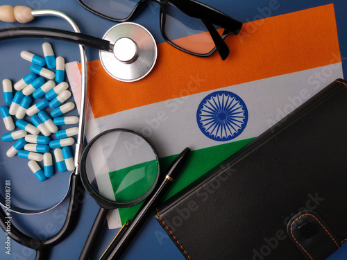 Medical tools, blue capsules, glasses, and a notebook surrounding the national flag of India on a blue surface