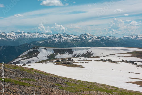 Foto Scenic alpine landscape with sunlit snowy field on stony hill with view to big mountain range with forest and snow far away under cloudy sky