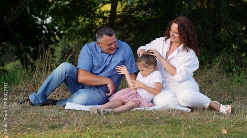 Happy mature family of three sits on blanket in summer park at sunset, laughing. Dad tickles daughter while Mom does her hair, then everyone tickles Mom. Family bonds, late fatherhood, recreation