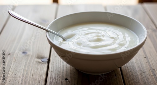White ceramic bowl filled with creamy yogurt and a silver spoon, rustic wooden table surface background.