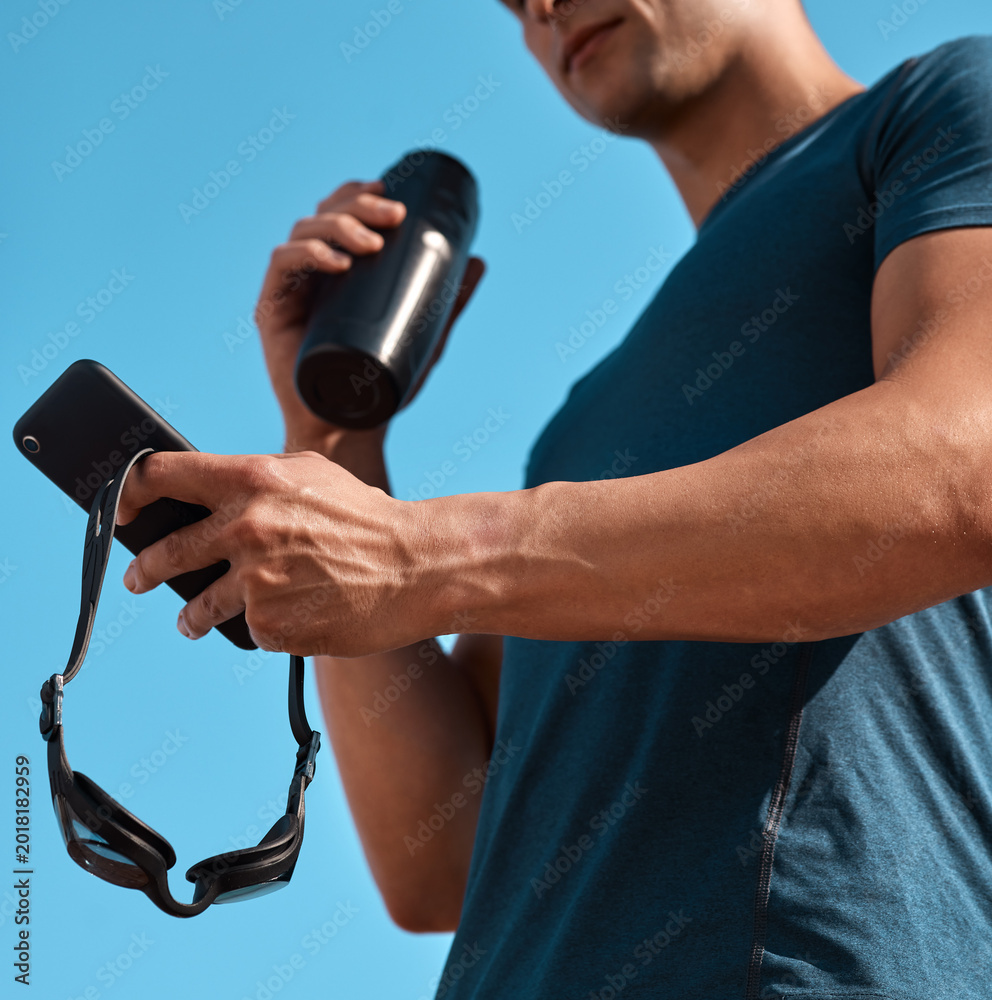 Obraz premium Hands, athlete and man with cellphone outdoor for texting, networking or contact at practice. Water bottle, fitness and male swimmer with phone for social media on mobile app at competition training.