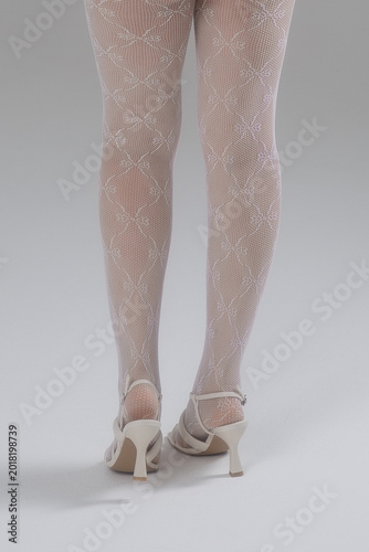 Beautiful female legs in white sandals and white lace tights on a gray background. Crossed women's legs in white pantyhose.