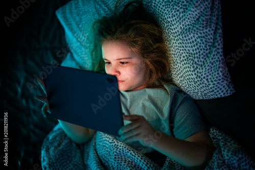 Kid facing tablet light in bed at night. Teenager lying in bed watching tablet at night.