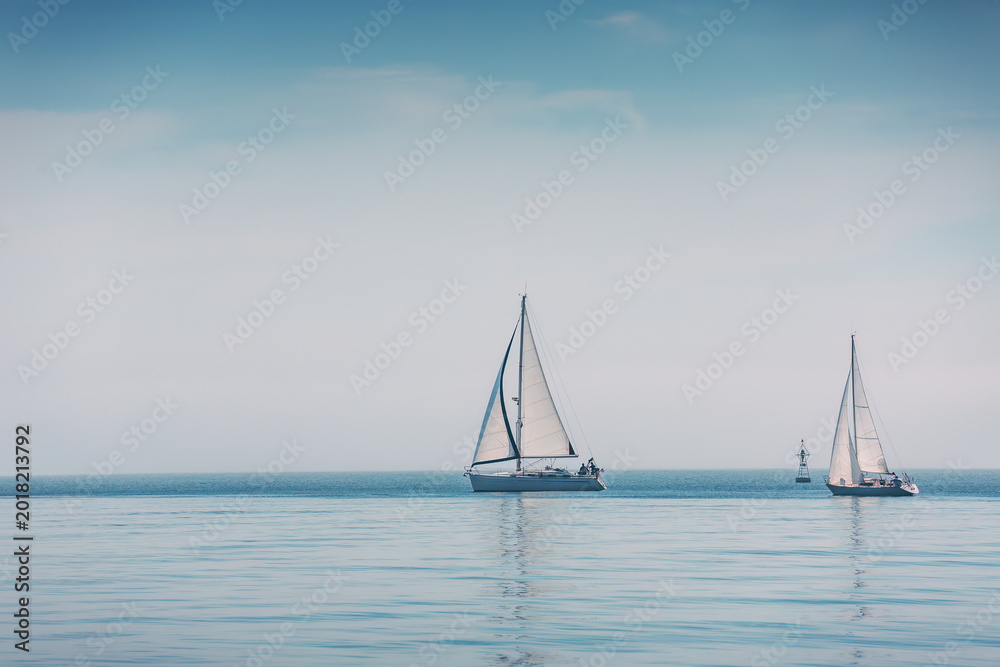 Naklejka premium Two Sailboats With White Sails and With Crew Aboard Sailing on a Calm Foggy Sea Under a Pale Blue Sky Near the Horizon