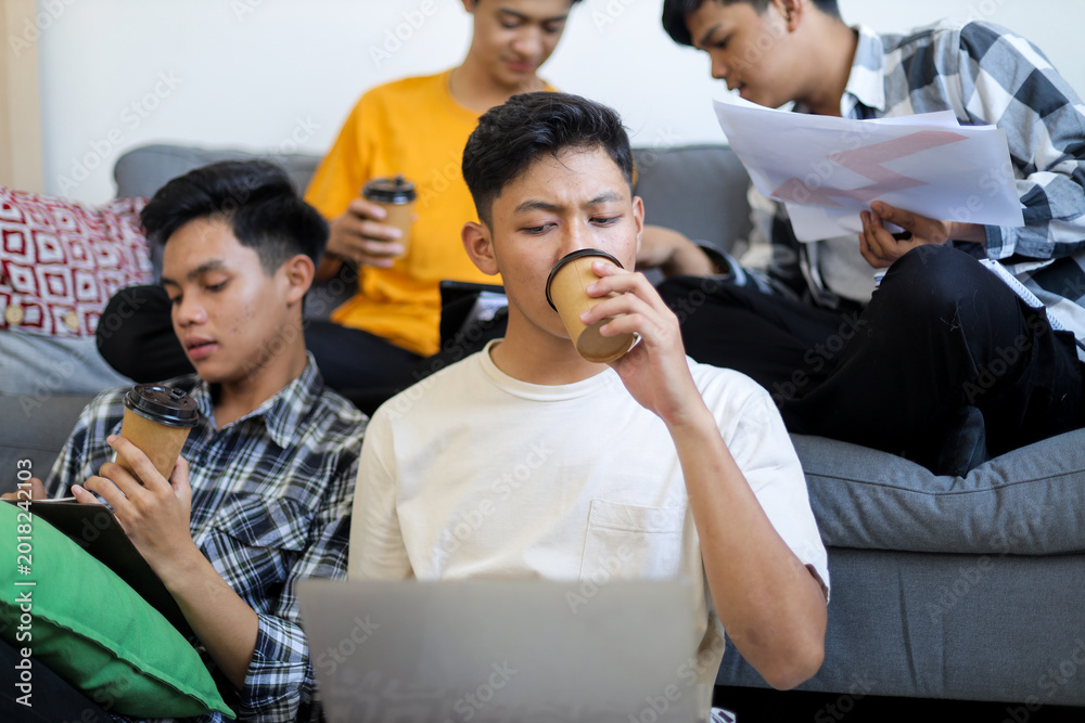 Fototapeta premium Four Young Asian Men Study Together in Cozy Living Room, Drinking Coffee and Using Laptop