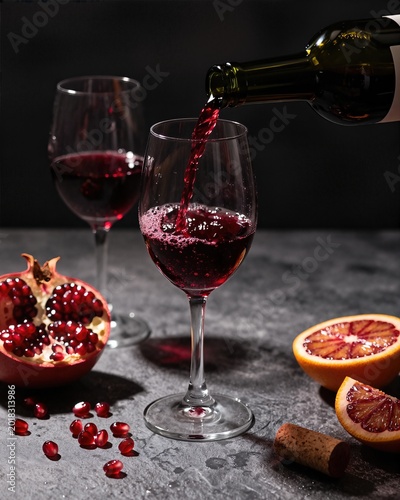 Red wine pours from a bottle into a glass. Pomegranate fruit halves sit on a dark stone table near a glass of red wine. Glass of red wine captures the light against a dark background.