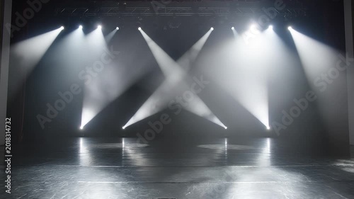 Empty performance stage with rustic wooden floorboards illuminated by moving white spotlights and smoke effects.