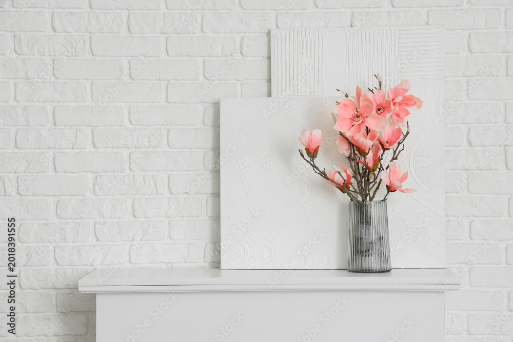 Fototapeta premium Vase with artificial magnolia flowers and paintings on fireplace near white brick wall