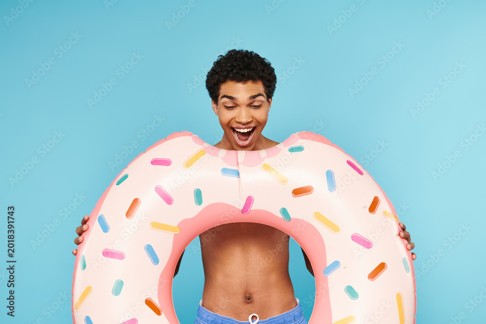 Obraz premium Joyful young man holds a giant donut float against a vibrant blue backdrop at summer pool party
