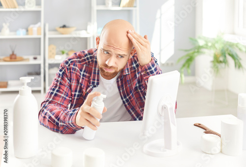 Bilde på lerret Young man in casual clothes looking in the mirror on his bald head sitting at table with cosmetics bottles at home preparing for beauty spa day