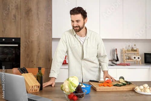 Handsome bearded man with laptop cooking in kitchen