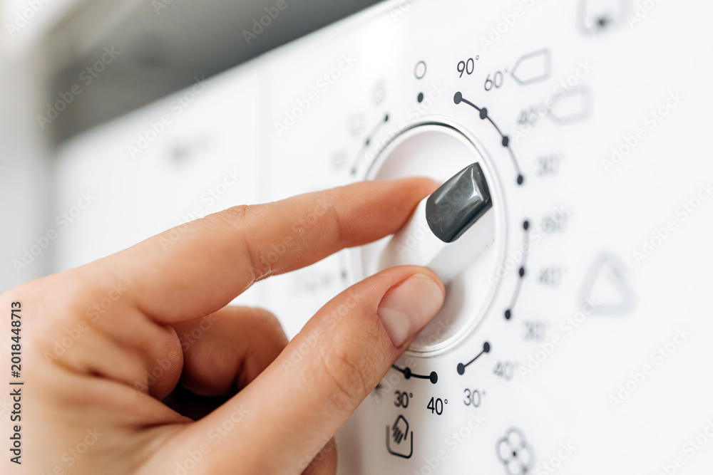 Fototapeta premium Hand adjusting the temperature dial on a modern washing machine with various settings and symbols visible on the control panel in a domestic laundry environment