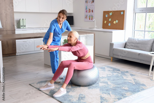 Female physiotherapist working with mature woman on fitball in rehabilitation...