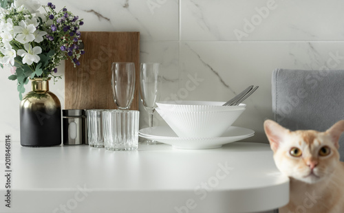 Tableau sur toile Minimal dining table setting with white dishes, flowers and blurred cat in bright modern apartment interior
