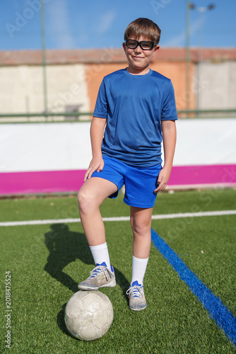 Young boy in sports glasses standing on a soccer pitch with foot on a football ball, actively training outdoors