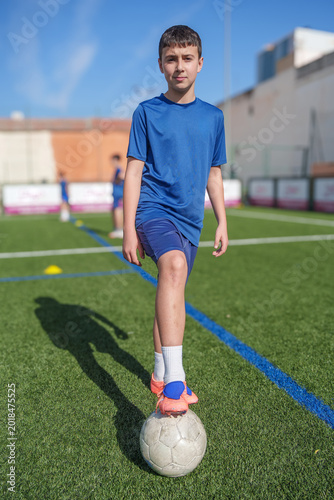 Young boy in a blue uniform standing on a soccer field, balancing a football on his foot during training