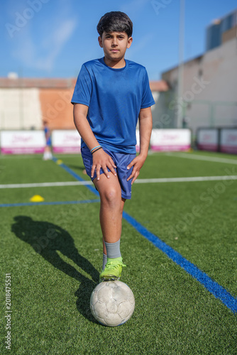 Boy standing confidently on a green artificial turf soccer field, foot resting on a ball, practicing for sport