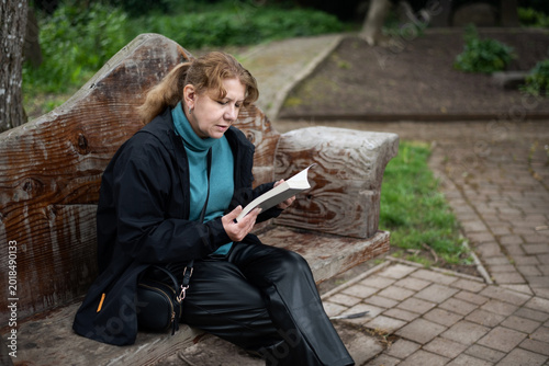 Photos A middle-aged woman sits on a carved wooden bench in a park, absorbed in reading a paper book