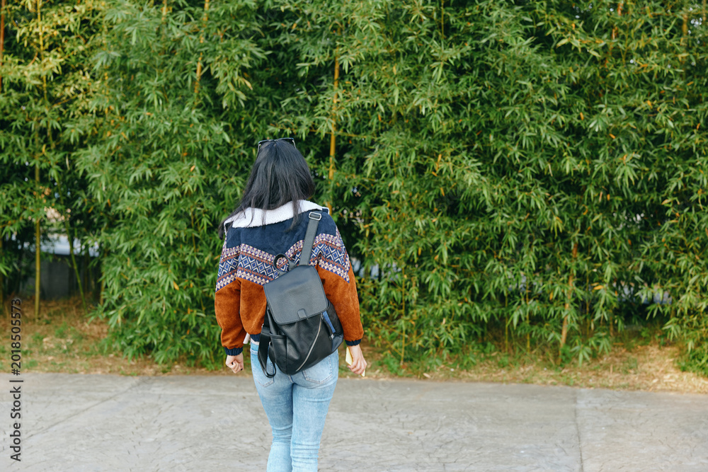 Obraz premium Back view of a woman with a backpack walking along a sidewalk among tall trees in casual autumn attire, conveying outdoor exploration and a calm park atmosphere