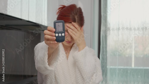 A woman in a kitchen displays a glucometer reading of 141 mg dL. This indicates a high blood sugar level, relevant for those monitoring diabetes.