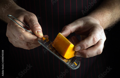 Hands are holding a cheese grater and a block of cheese as it is being grated. The setting is a kitchen with low light. The focus is on the action of grating