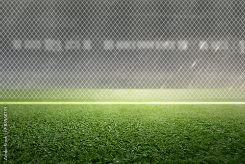 Close up of green artificial turf on a sports field with bright stadium lights and a chain-link fence in the blurred background. Ideal for soccer, football, and athletic concepts