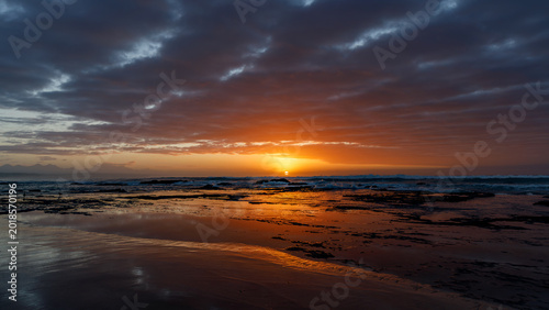 Aerial view of the sun setting over the rocky coastline of Hartenbos beach with dramatic clouds and golden reflections on the water Mossel Bay, Western Cape, South Africa.