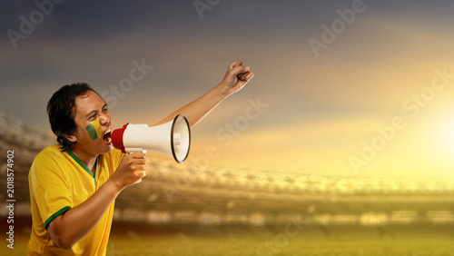 An enthusiastic Indonesian southeast asian soccer supporter with face paint shouting into a megaphone and raising a fist in celebration at a sports stadium during sunset