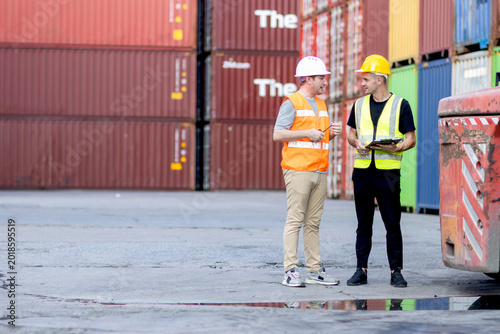 Two workers with safety vest and helmet working together at logistic shipping cargo container yard. Male worker writing on clipboard during discussing with his engineer foreman who holding worky talky