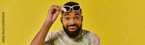 Fotografía Handsome young man joyfully adjusting sunglasses against a vibrant yellow backgr