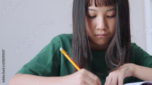Close up selective focus of a concentrated Asian kid, a young Thai girl or Chinese student, diligently doing homework and writing with a pencil in a notebook. Education, learning, and study concept.