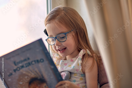 Принти на полотні Happy little girl in blue glasses reading a colorful children’s book by the window at home