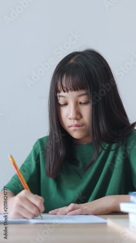 Vertical video close up and selective focus showing Asian kid girl concentrating as she writes in her notebook with a pencil. Bright daylight fills the room while she is doing her homework.