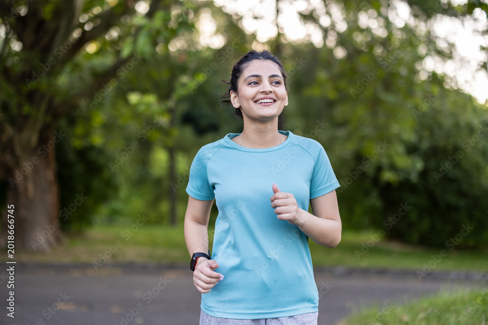 Fototapeta premium Young indian woman feeling positive and healthy while running outdoors, wearing sportswear and a smartwatch, focusing on her fitness goals in a green park environment