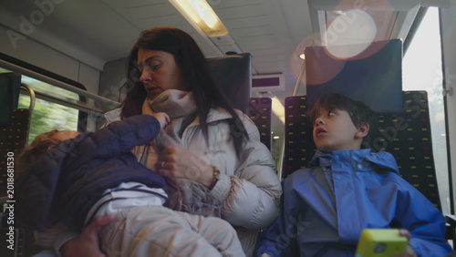 Принти на полотні Mother sits on train holding restless toddler while older child looks upward cre
