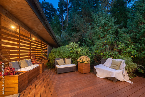 A wooden terrace with an outdoor area surrounded by lush greenery. Wicker garden furniture. Summer evening.