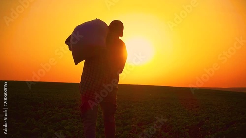 Silhouette of a male farmer with a sack of wheat on his shoulder. A male worker at sunset with a sack of fertilizer in his hand. The concept of farming, agriculture, hard work of workers.
