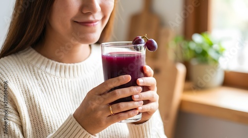 Smiling young woman holding a glass of vibrant purple grape juice garnished with fresh grapes, enjoying a healthy drink in a cozy home setting with warm natural light.