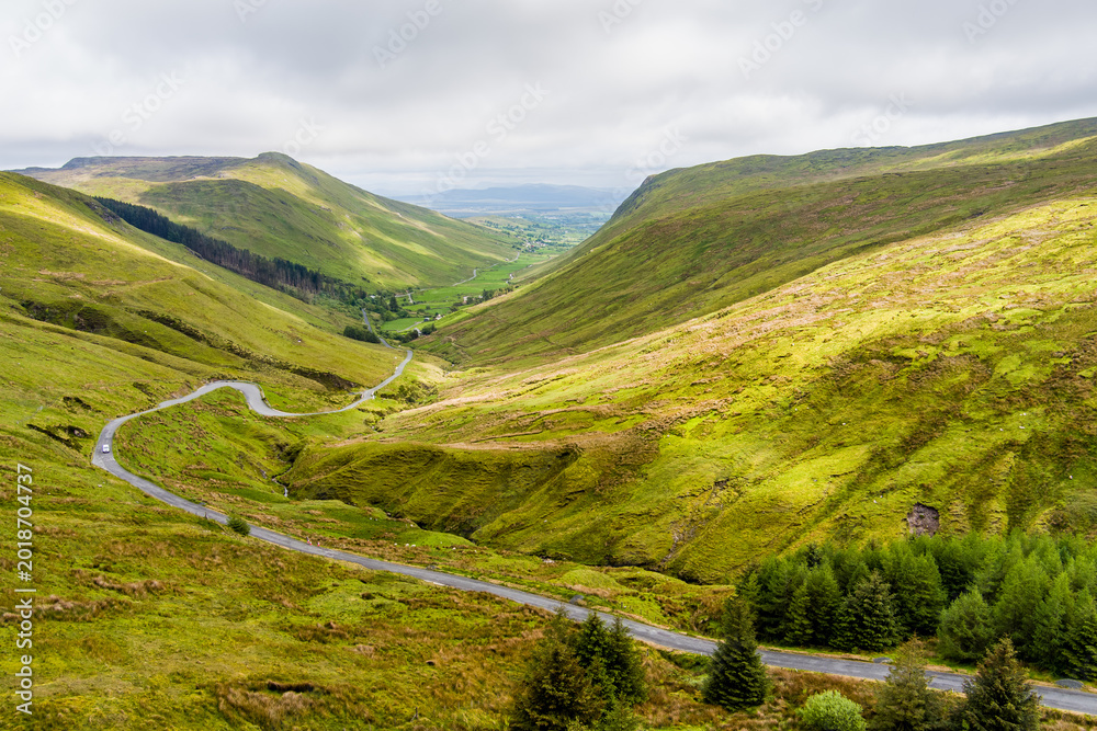Naklejka premium Aerial view of Glengesh Pass curving through a broad green valley in County Donegal, Ireland. A dramatic birds eye landscape of remote mountains and winding roads