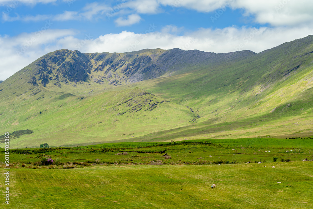 Obraz premium Sunlit mountains and green fields spread across the landscape near Killary Fjord in western Ireland. A broad rural panorama of the Connemara and Mayo border region.