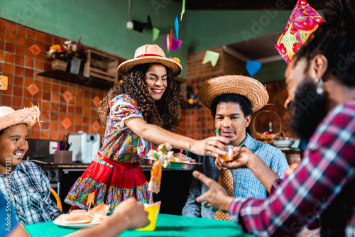 Happy Brazilian friends and family celebrating Festa Junina with traditional food and drinks, wearing straw hats and colorful costumes during the June Festival celebration.
