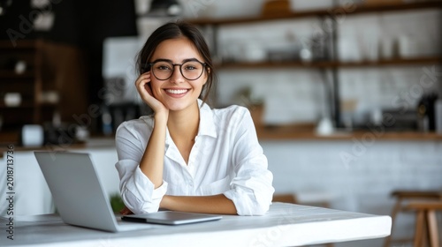 Young woman with glasses smiles warmly, working on a laptop at a light-filled table in a cafe