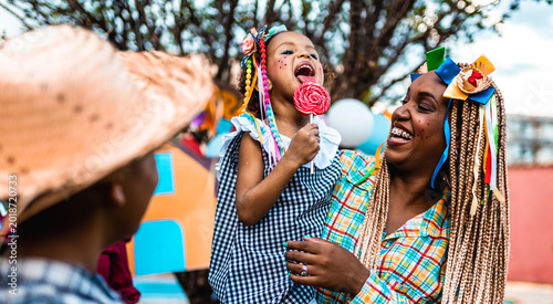 Happy Afro-Brazilian mother and daughter celebrating a traditional Festa Junina festival with a large lollipop.