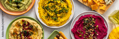 A variety of hummus flavors including turmeric, classic, and broccoli are served with fresh bread, nuts, and garnishes on a colorful charcuterie board, perfect for sharing, banner
