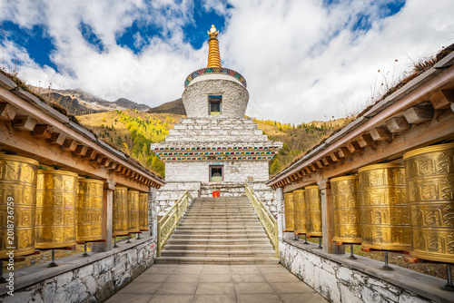 Fotografie A magnificent white Tibetan Buddhist stupa flanked by rows of large golden prayer wheels stands at Siguniangshan (Four Sisters Mountain) National Park in Xiaojin County, Sichuan Province, China
