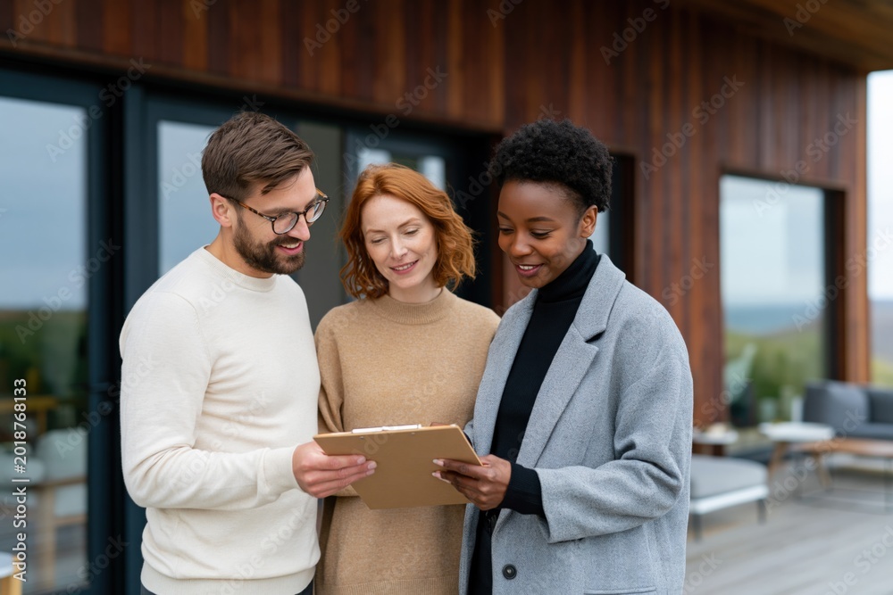 Fototapeta premium Diverse real estate team discussing contract and floorplans with young couple in front of modern wooden house for home tour and client meeting