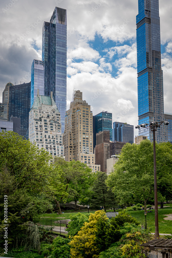 Fototapeta premium Central Park Trees Below Midtown Manhattan Towers. Central Park greenery with footpaths, lamps, and Midtown Manhattan skyscrapers under cloudy sky.